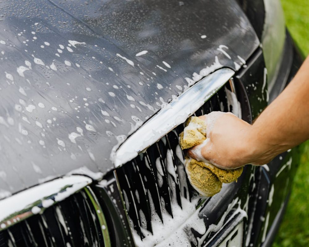 Detailed view of a hand washing a car grille with a soapy sponge outdoors.