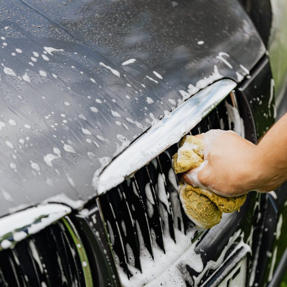 Detailed view of a hand washing a car grille with a soapy sponge outdoors.